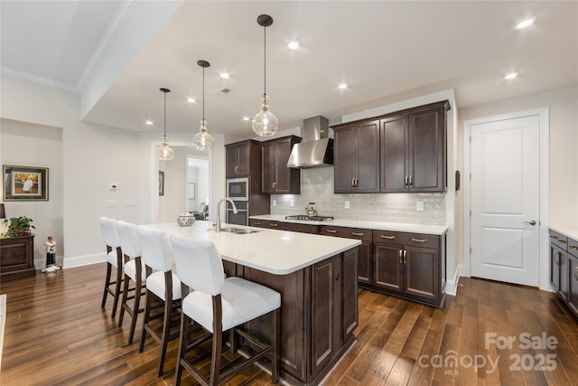 kitchen featuring wall chimney exhaust hood, sink, hanging light fixtures, appliances with stainless steel finishes, and an island with sink