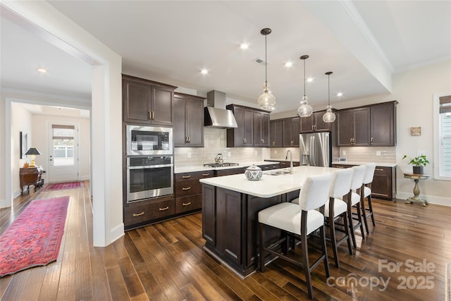 kitchen with sink, hanging light fixtures, a kitchen island with sink, stainless steel appliances, and wall chimney exhaust hood