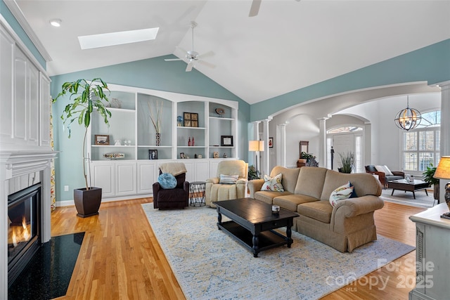 living room featuring vaulted ceiling, light wood-type flooring, built in shelves, ceiling fan with notable chandelier, and ornate columns