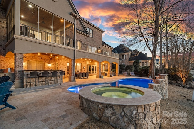 pool at dusk with an in ground hot tub, an outdoor bar, and a patio