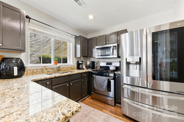 kitchen with light stone countertops, appliances with stainless steel finishes, light wood-type flooring, dark brown cabinetry, and sink