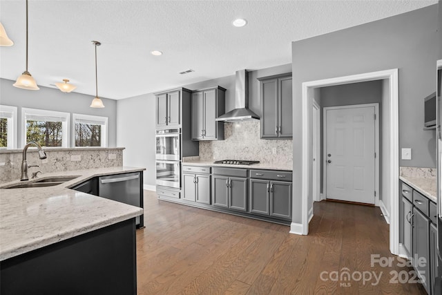 kitchen with decorative light fixtures, wall chimney range hood, sink, appliances with stainless steel finishes, and light stone counters