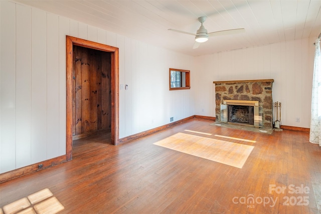 unfurnished living room featuring wood-type flooring, ceiling fan, a stone fireplace, and baseboards