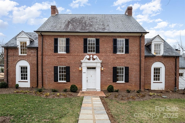 view of front facade featuring brick siding, a chimney, a front lawn, and a high end roof