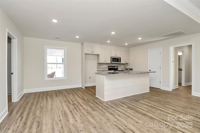 kitchen featuring white cabinetry, an island with sink, appliances with stainless steel finishes, light wood-type flooring, and light stone counters