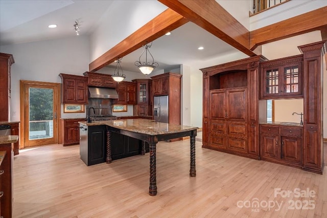 kitchen featuring pendant lighting, stainless steel fridge, range hood, a center island with sink, and beamed ceiling