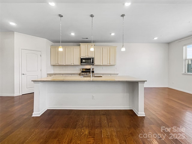 kitchen featuring pendant lighting, appliances with stainless steel finishes, dark wood-type flooring, cream cabinets, and a kitchen island with sink