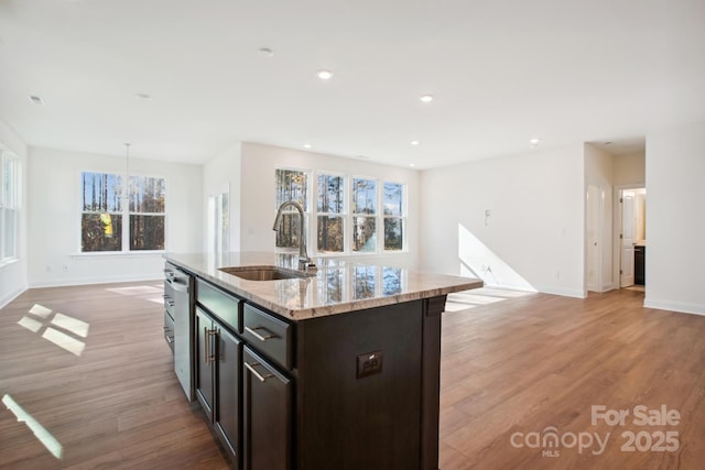 kitchen with light stone countertops, dishwasher, sink, dark brown cabinets, and an island with sink