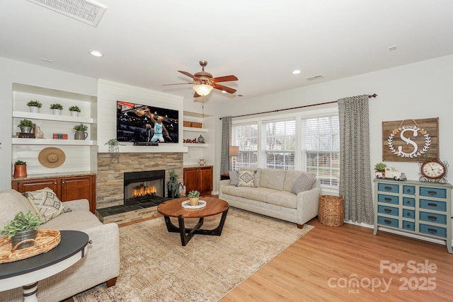 living room with ceiling fan, a stone fireplace, built in features, and light wood-type flooring