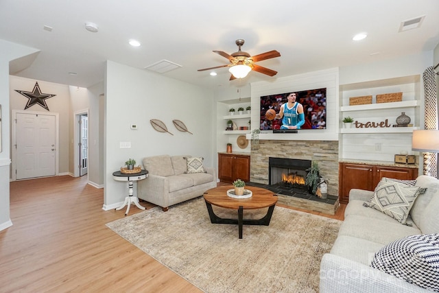 living room featuring built in shelves, a stone fireplace, ceiling fan, and light hardwood / wood-style flooring