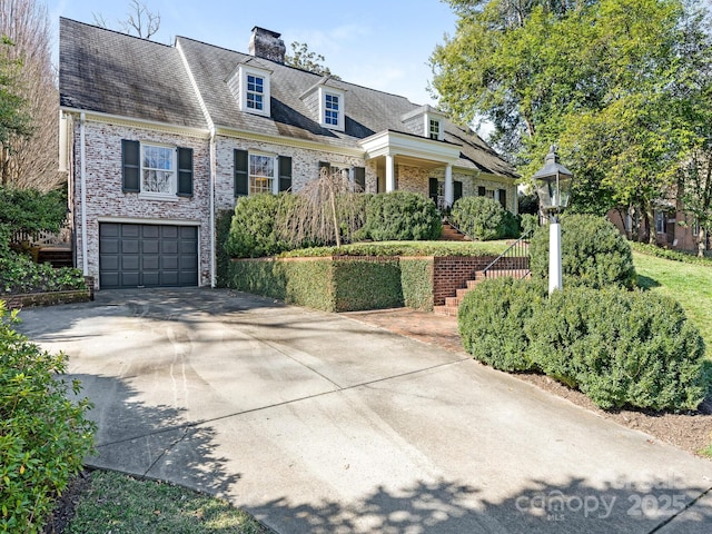 new england style home with an attached garage, brick siding, concrete driveway, stairway, and a chimney