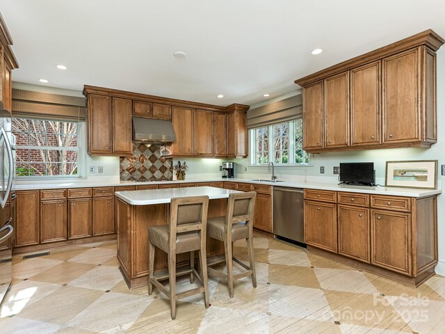 kitchen with under cabinet range hood, a sink, stainless steel dishwasher, a center island, and brown cabinetry