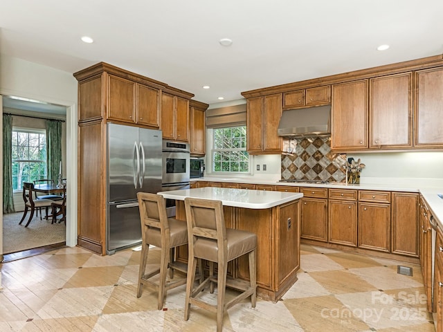 kitchen featuring under cabinet range hood, a kitchen island, stainless steel appliances, and brown cabinetry