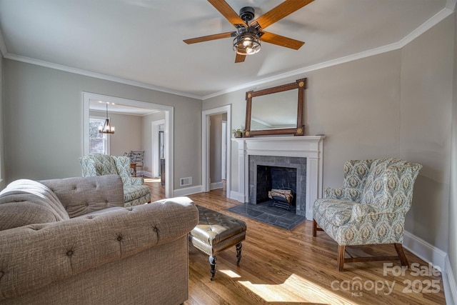 living room featuring crown molding, wood-type flooring, a tile fireplace, and ceiling fan with notable chandelier