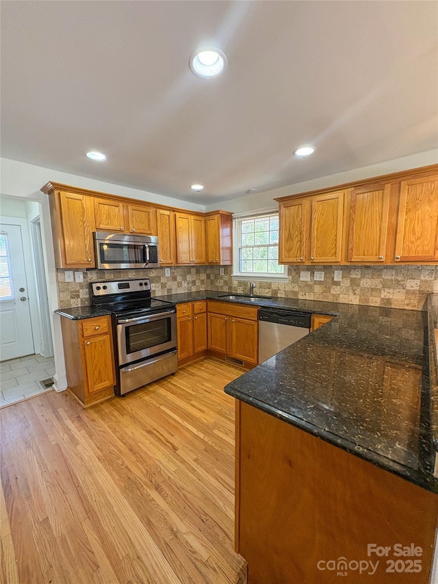 kitchen with appliances with stainless steel finishes, brown cabinets, dark stone countertops, light wood-type flooring, and backsplash