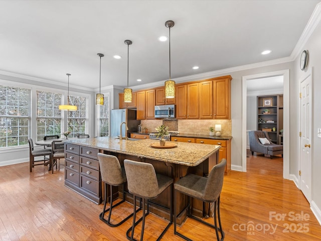 kitchen with crown molding, wood-type flooring, appliances with stainless steel finishes, an island with sink, and light stone countertops