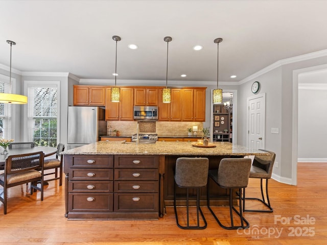 kitchen with appliances with stainless steel finishes, an island with sink, light stone counters, crown molding, and light hardwood / wood-style flooring