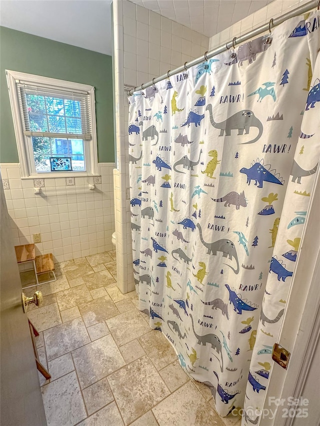 bathroom featuring wainscoting, a shower with shower curtain, stone tile flooring, and tile walls