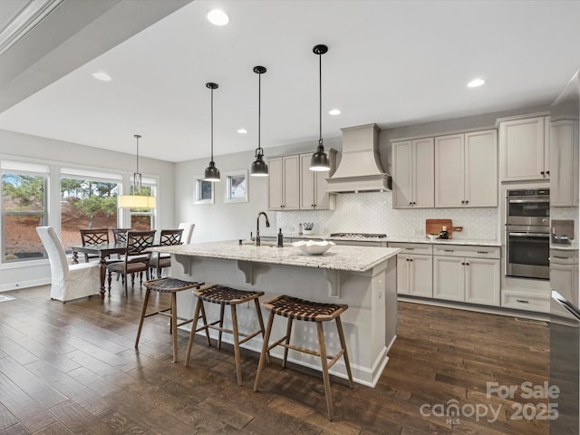 kitchen with appliances with stainless steel finishes, pendant lighting, an island with sink, custom exhaust hood, and light stone countertops