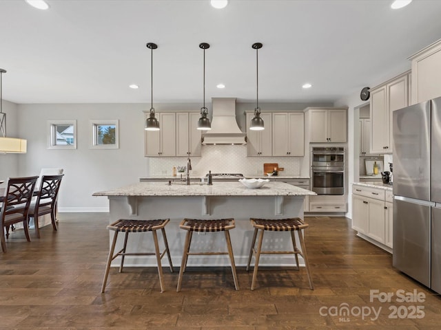 kitchen featuring decorative light fixtures, stainless steel appliances, custom range hood, and an island with sink