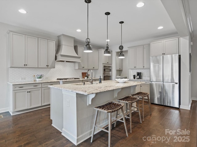 kitchen with a kitchen island with sink, hanging light fixtures, stainless steel appliances, light stone counters, and custom exhaust hood