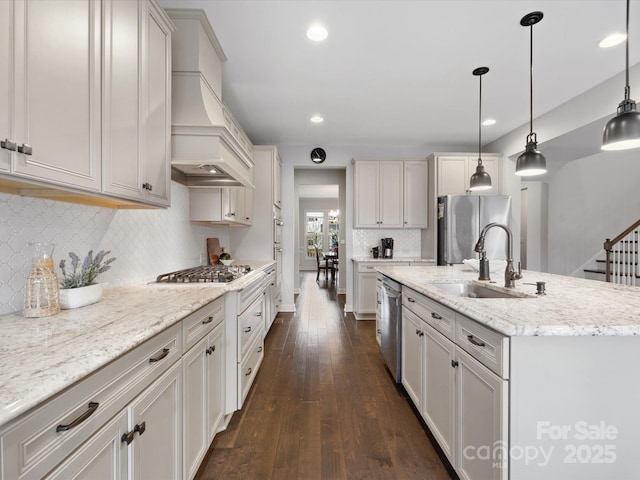 kitchen with decorative light fixtures, sink, stainless steel appliances, custom range hood, and a center island with sink