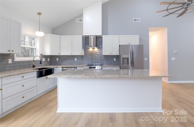 kitchen featuring appliances with stainless steel finishes, white cabinetry, wall chimney range hood, decorative light fixtures, and a kitchen island