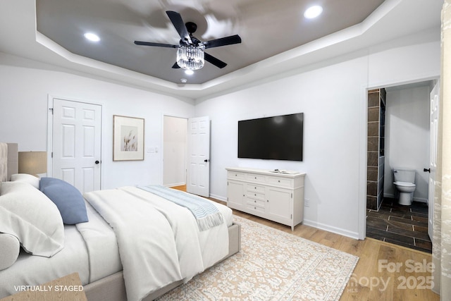 bedroom with ceiling fan, light hardwood / wood-style flooring, ensuite bath, and a tray ceiling