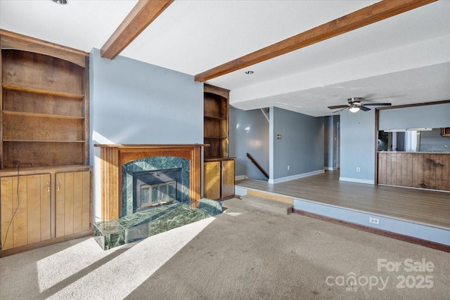 unfurnished living room with a premium fireplace, ceiling fan, beam ceiling, carpet floors, and built in shelves