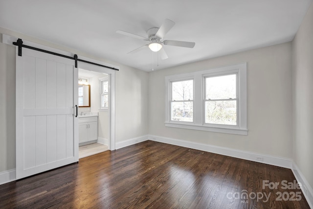 unfurnished bedroom with ceiling fan, a barn door, dark wood-type flooring, and ensuite bath