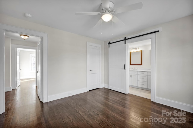 unfurnished bedroom featuring ensuite bathroom, dark hardwood / wood-style flooring, sink, ceiling fan, and a barn door