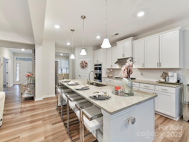 kitchen with white cabinetry, decorative light fixtures, stainless steel appliances, an island with sink, and light stone countertops