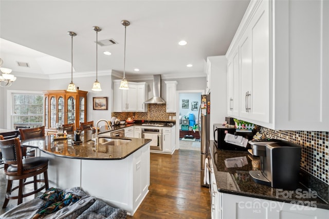 kitchen with wall chimney range hood, sink, appliances with stainless steel finishes, hanging light fixtures, and white cabinets