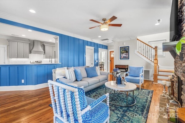 living room with a stone fireplace, sink, crown molding, light wood-type flooring, and ceiling fan