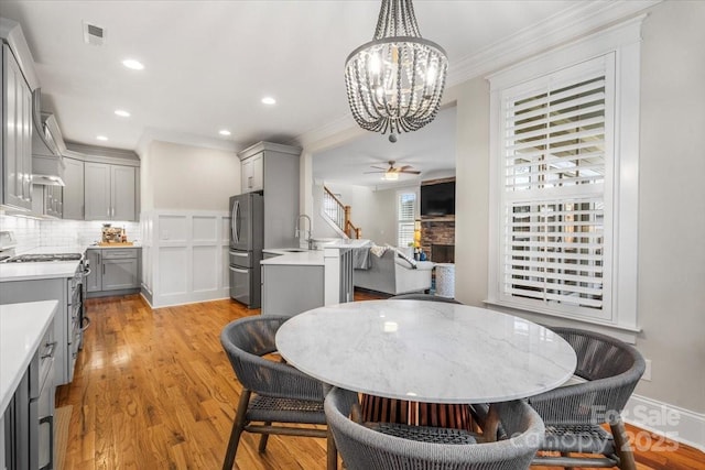 dining room with a stone fireplace, ceiling fan with notable chandelier, sink, ornamental molding, and light hardwood / wood-style flooring