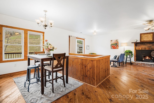 dining area featuring a brick fireplace, ceiling fan with notable chandelier, and light wood-type flooring