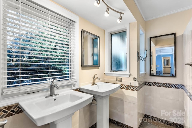 bathroom with ornamental molding, two sinks, wainscoting, and tile walls