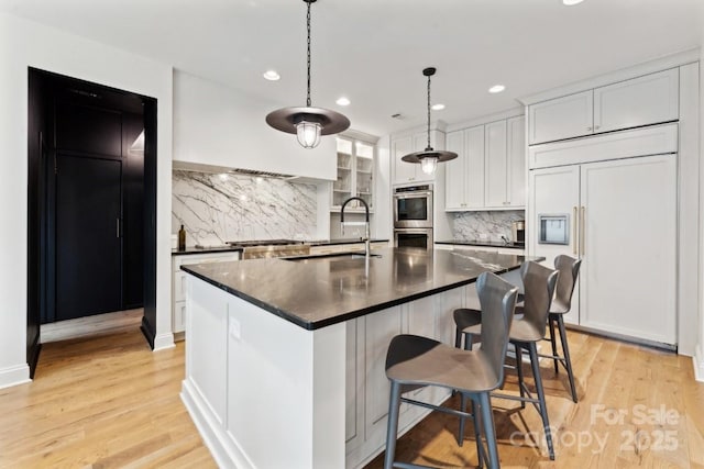 kitchen featuring white cabinets, dark countertops, an island with sink, hanging light fixtures, and paneled built in refrigerator