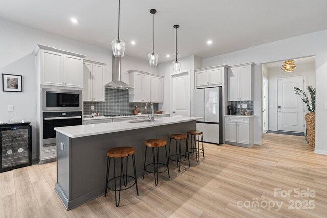 kitchen featuring wall chimney exhaust hood, stainless steel appliances, an island with sink, white cabinets, and sink