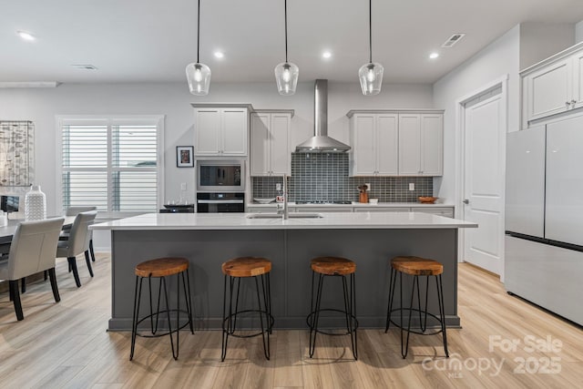 kitchen with appliances with stainless steel finishes, sink, wall chimney range hood, hanging light fixtures, and a kitchen island with sink