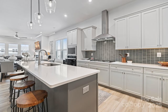 kitchen with appliances with stainless steel finishes, wall chimney range hood, decorative light fixtures, sink, and a kitchen island with sink