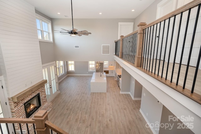 living room featuring a towering ceiling, light hardwood / wood-style flooring, ceiling fan, a brick fireplace, and french doors