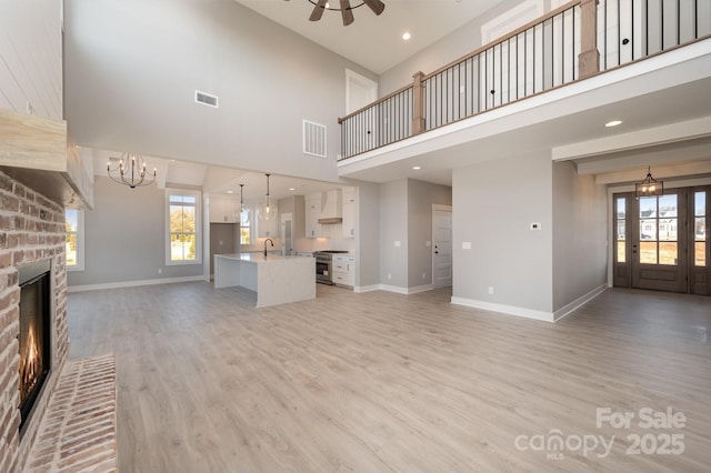 unfurnished living room featuring sink, a brick fireplace, light wood-type flooring, a towering ceiling, and ceiling fan with notable chandelier