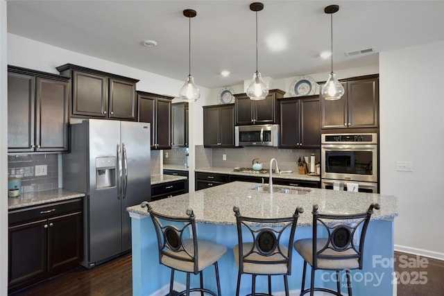 kitchen with a center island with sink, a kitchen breakfast bar, stainless steel appliances, light stone counters, and decorative light fixtures