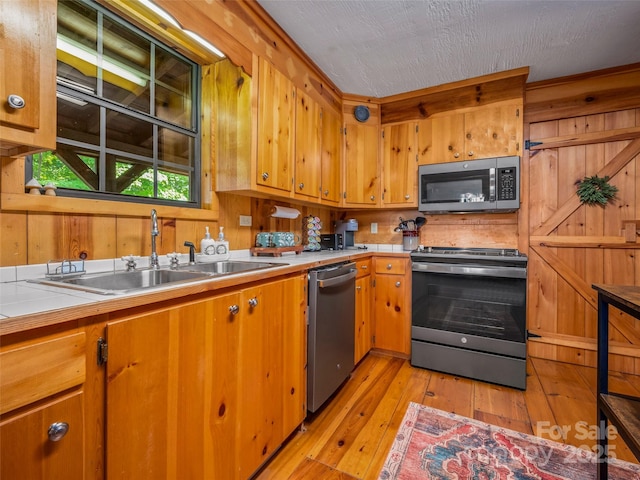 kitchen with a textured ceiling, wooden walls, sink, light hardwood / wood-style flooring, and stainless steel appliances