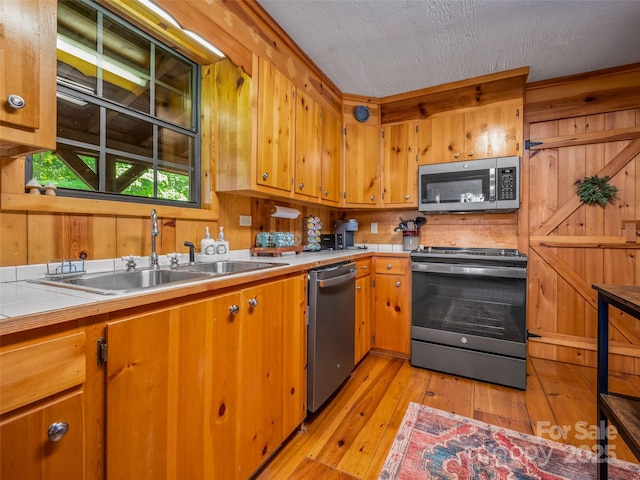 kitchen with appliances with stainless steel finishes, sink, a textured ceiling, wood walls, and light hardwood / wood-style floors