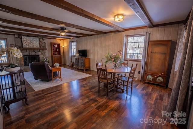 dining room with ceiling fan, a stone fireplace, beam ceiling, and dark hardwood / wood-style floors