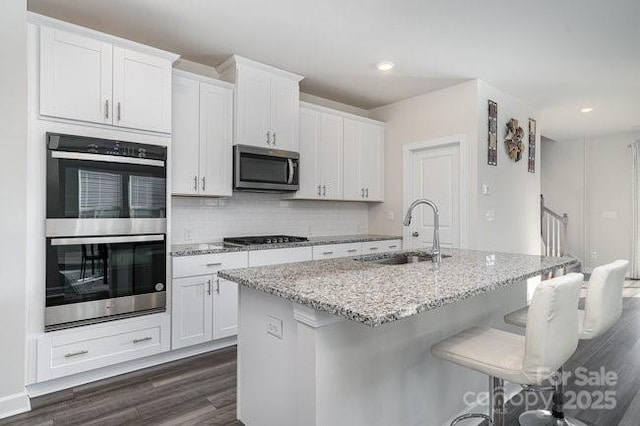 kitchen with appliances with stainless steel finishes, white cabinetry, sink, light stone counters, and a center island with sink