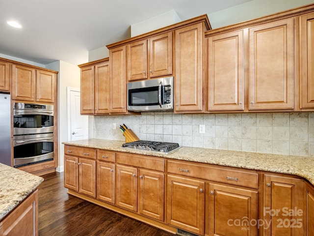 kitchen with light stone countertops, stainless steel appliances, backsplash, brown cabinets, and dark wood-style floors