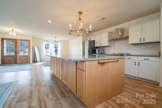 kitchen with a center island with sink, stainless steel refrigerator, white cabinetry, a chandelier, and wall chimney exhaust hood